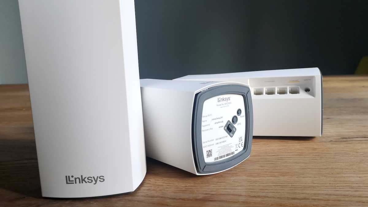 Three Linksys Atlas 6 units on a table, one stood up, facing forwards, the other two laying flat, showing the rubberised feet and Ethernet ports, respectively